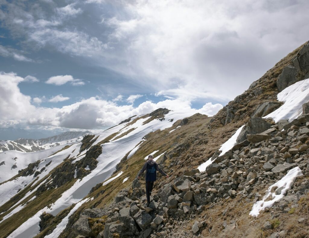 person in blue jacket and black pants standing on rocky mountain under white clouds during daytime